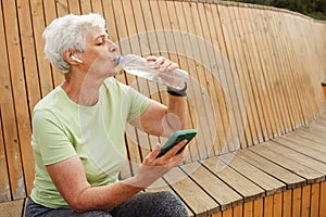 An elderly happy pensioner woman after a run sits on a wooden bench and drinks water.