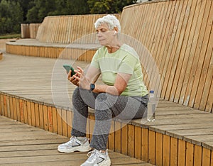 An elderly happy retired woman after a run sits on a wooden bench and sends a text message.