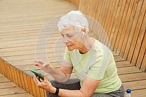 An elderly happy retired woman after a run sits on a wooden bench and sends a text message.