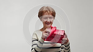 An elderly happy woman catches, says wow, holds in hands, considers and cuddles with amazement a bright crimson gift box