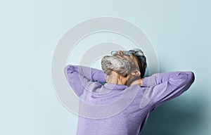 Gray-haired man in purple sweater, sunglasses. Put his hands behind head and looking up, posing on blue background. Close up