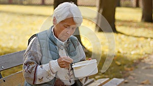 Elderly homeless retired caucasian woman sitting on a bench outdoors and eating warm soup that was given to her by a