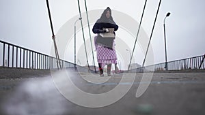 Elderly homeless woman stay on the bridge near the river port in cold windy grey weather asking for help and begging