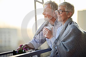 An elderly hugged couple in love is enjoying a view on the city from the terrace of their apartment at a beautiful sunset.