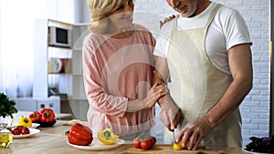 Elderly husband cutting fresh pepper in kitchen, wife hugging love, closeness