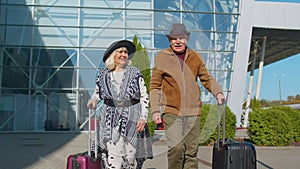 Elderly husband wife retirees tourists go to airport terminal for boarding with luggage on wheels