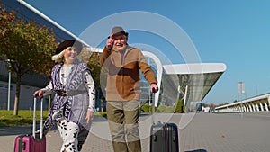 Elderly husband wife retirees tourists go to airport terminal for boarding with luggage on wheels