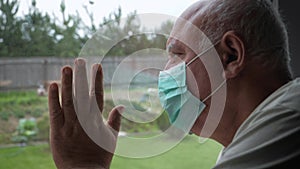 Elderly Illness Man In Medical Mask Looks Out Window And Puts His Hand On Glass