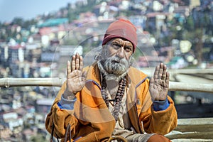 Elderly Indian beard man, two hands open, glance forward, wearing cultural rope and beads with walking stick.