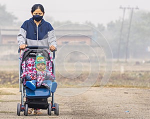 an elderly indian lady wearing black face mask pushing an indian baby in winter wear in a stroller perambulator on a road with