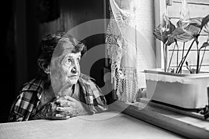 An elderly lady sits sadly near the window of his old house.