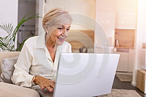 An elderly lady using a laptop. Portrait of beautiful older woman working laptop computer indoors. Senior woman sitting at sofa