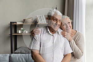 Elderly loving couple standing in living room looking out window