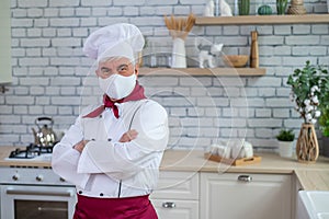 An elderly male chef wearing a mask in the kitchen crossed his arms over chest