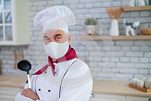 An elderly male chef wearing a mask in the kitchen crossed his arms over chest