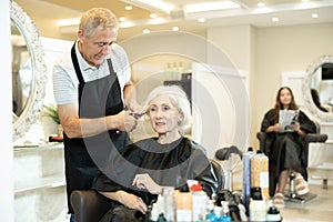 Elderly male hairdresser performs hair styling and creates hairstyle with curling iron and comb