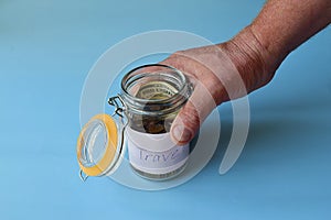 Elderly male hand holds a glass jar with money savings, banknotes and coins on a blue background in sunlight. Travel money savings