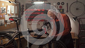 Elderly male mechanic working in bicycle repair shop or garage, cleaning wheel with rag while repairing bike