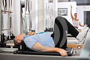 Elderly male pensioner lying down on bed of Pilates reformer bed machine with legs up, working out and recovering in gym