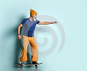 Elderly male in t-shirt, orange pants, hat, gumshoes. Riding black skateboard posing sideways on blue background. Full length