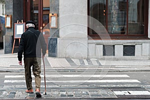 An elderly male with a walking stick slowly walks across a crosswalk in the middle of a city alone
