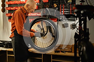 Elderly male worker holding and repairing bicycle wheel while standing in authentic bicycle workshop.