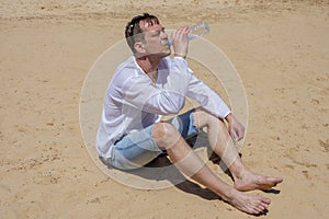 An elderly man 45-50 years old greedily drinks water from a plastic bottle sitting on the sand in the desert. Concept: thirst and