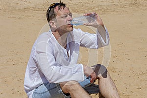 An elderly man 45-50 years old greedily drinks water from a plastic bottle sitting on the sand in the desert. Concept: thirst and