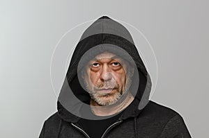 An elderly man 50-55 years old with a beard and wearing a black hooded hoodie looks into the camera against