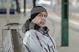 An elderly man 50-55 years old with a gray beard and wearing a hat sits on a metal bench at the train station.