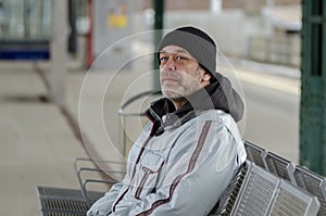 An elderly man 50-55 years old with a gray beard and wearing a hat sits on a metal bench at the train station.