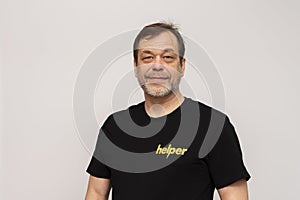 Elderly man 50-55 years old wearing a black T-shirt that says: Helper. Looking into the camera on a white background.