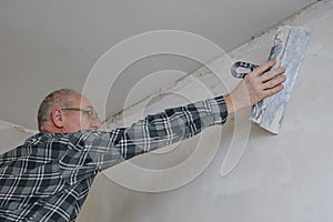 Elderly man 59 years old in a plaid shirt, glasses does the finishing of a plastered wall with a hand tool spatula in