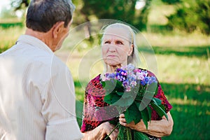 An elderly man of 80 years old gives flowers to his wife in a summer park.