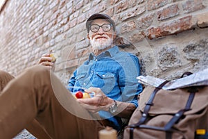 Elderly man with backpack sitting by brick wall, eating healthy homemade snack, resting. Traveling and solo trips in