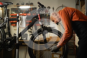 Elderly man bike mechanic repairs bicycle in workshop or garage. Portrait of man wearing apron fixes cycle wheel. Bike