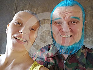 elderly man with blue beard and girl gray hair are standing next to each other, posing for camera and filming themselves on phone