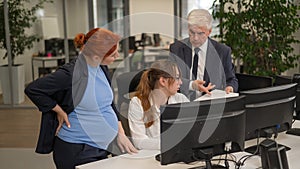 An elderly man, a Caucasian woman and a pregnant woman are discussing work issues at the computer.