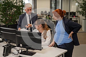 An elderly man, a Caucasian woman and a pregnant woman are discussing work issues at the computer.