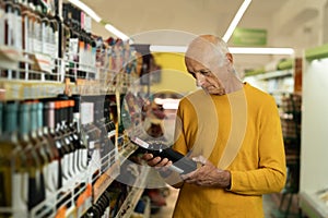 Elderly man chooses wine in the store. Senior man reads the label on a bottle of wine before buying