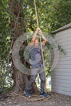 Elderly Man Climbing a Rope Hanging from a Tree in the Back Yard.