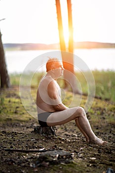 An elderly man without clothes is sitting on the lake shore
