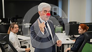 An elderly man with a clown nose stands next to two Caucasian women in the office.