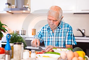 Elderly man cuts vegetables for salad at the table in the kitchen