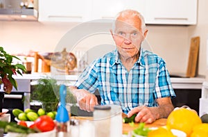 elderly man cuts vegetables for salad at the table in the kitchen