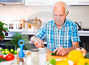 Elderly man cuts vegetables for salad at the table in the kitchen