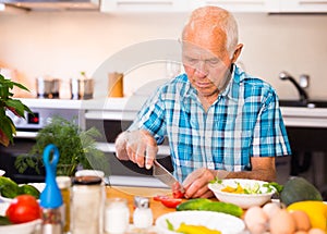 Elderly man cuts vegetables for salad at the table in the kitchen