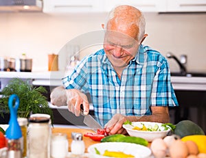 elderly man cuts vegetables for salad at the table in the kitchen