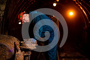 An elderly man dressed in work overalls and a helmet stands near the old inverted vogonetki. Miner