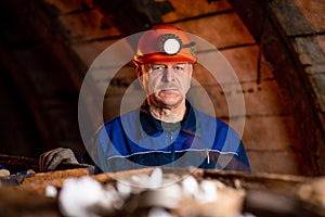 An elderly man dressed in work overalls and a helmet stands near the old inverted vogonetki. Miner